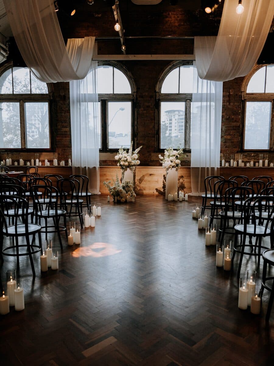 A ceremony space with arched windows, draped fabric and candles lining the aisle